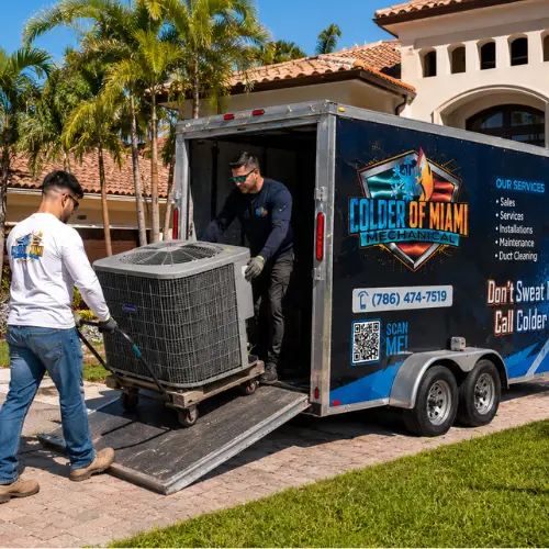 Two Colder of Miami HVAC technicians unloading a replacement air conditioning condenser unit from a service trailer at a residential driveway in Miami Beach.
